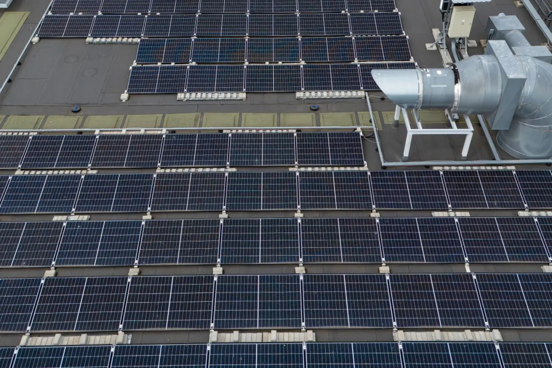 Aerial view of rows of solar modules on a flat commercial roof beside a rooftop exhaust unit